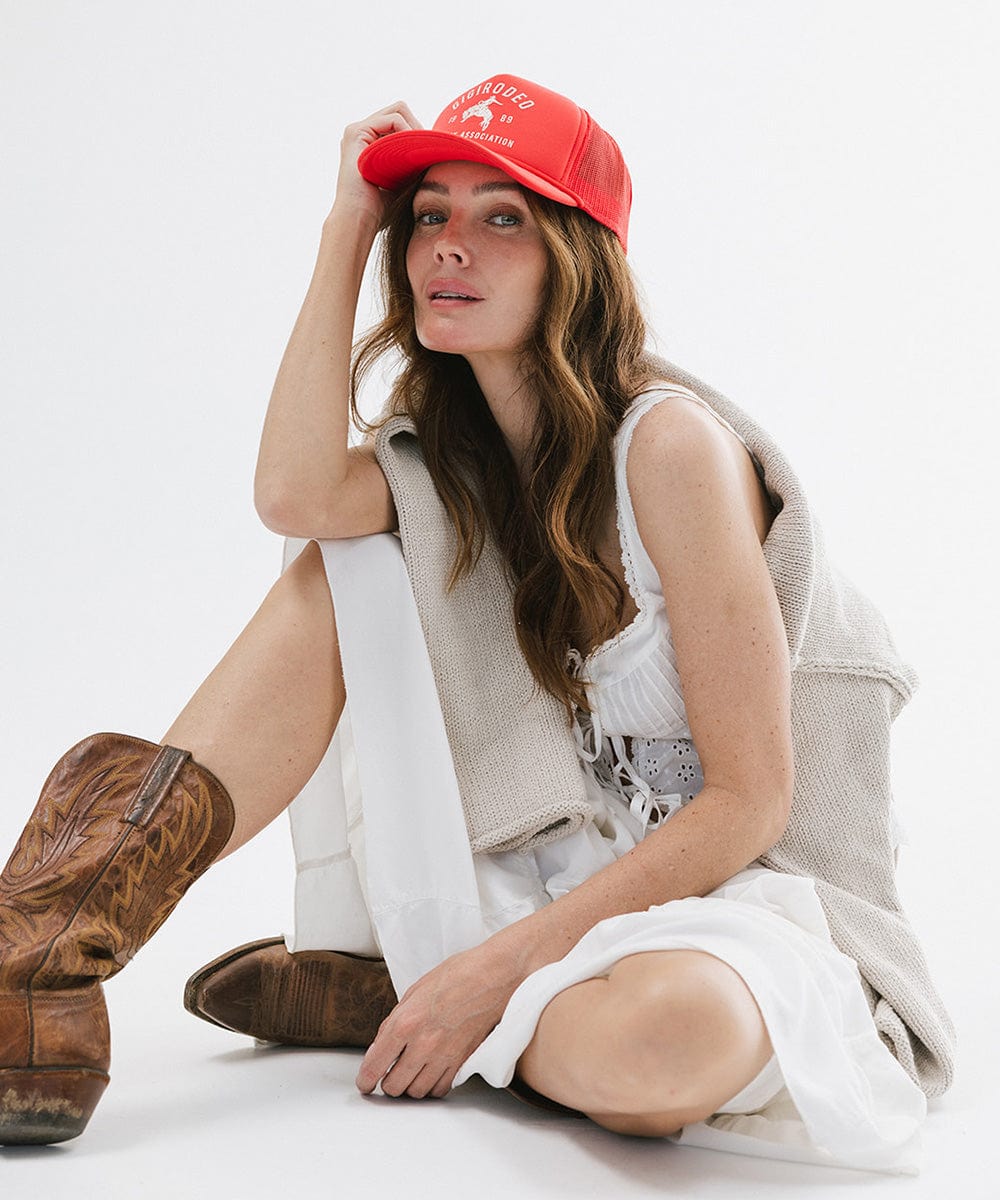 Woman wearing a vintage red cap with golden 'GIGIRODEO' text and logo, and a grey sweater, white pants and brown boots, sitting against a plain background #color_vintage red
