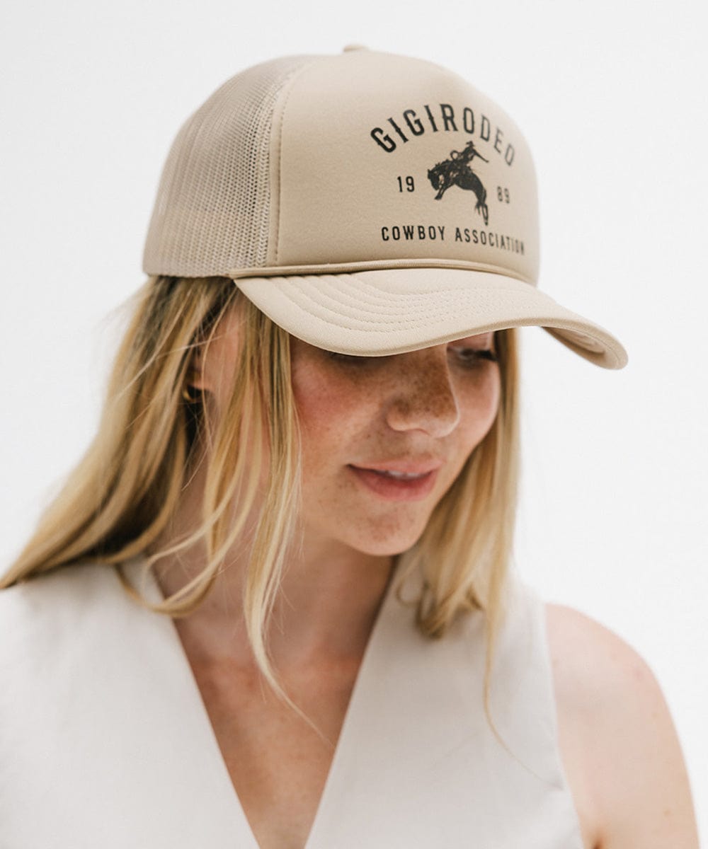 Woman wearing a tan cap with golden 'GIGIRODEO' text and logo, and a white top against a plain background #color_tan