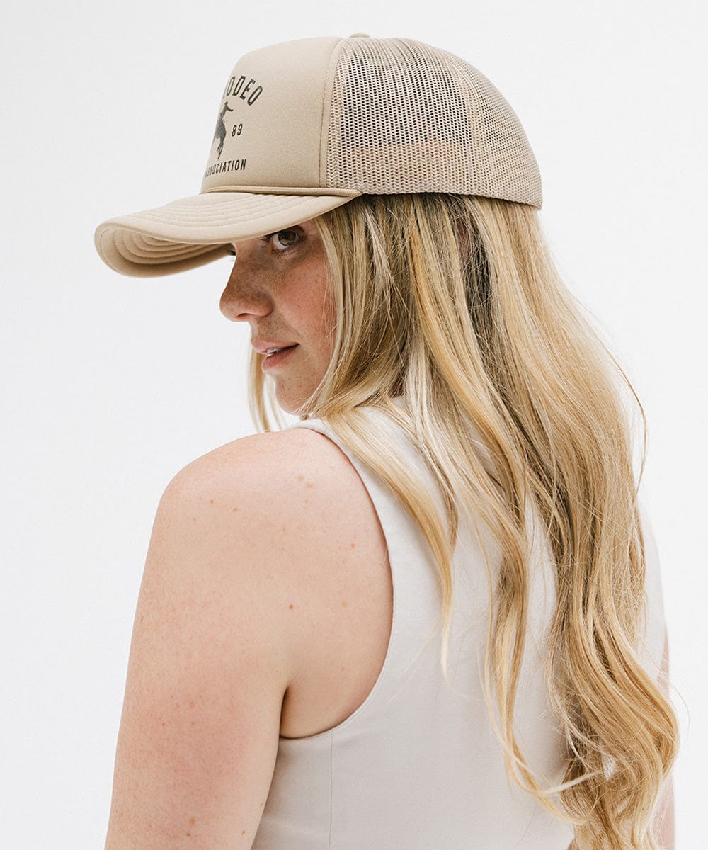 Woman wearing a tan cap with golden 'GIGIRODEO' text and logo, and a white top, facing sideways against a plain background #color_tan