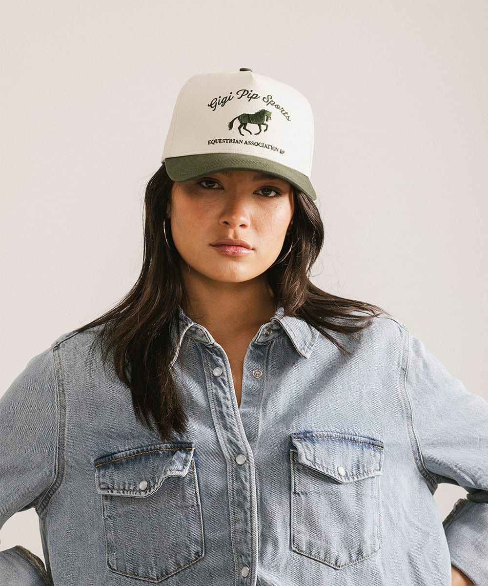 Woman wearing a cream and dark green Trucker hat with 'Gigi Pip Sports' and horse logo and a denim shirt against a plain background #color_cream - dark green