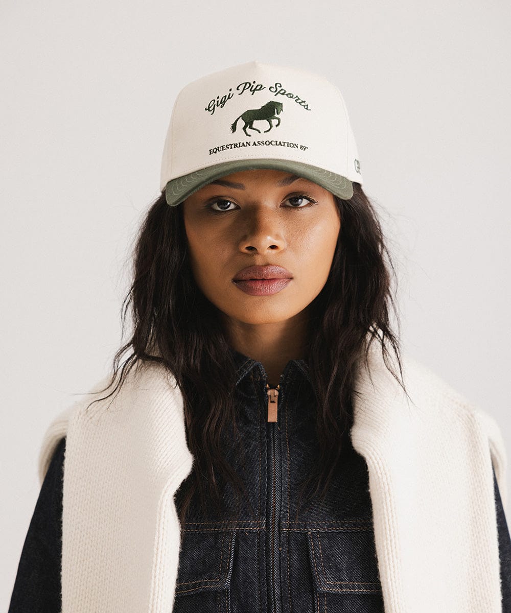Woman wearing a cream and dark green Trucker hat with 'Gigi Pip Sports' and horse logo and a denim jacket against a plain background #color_cream - dark green