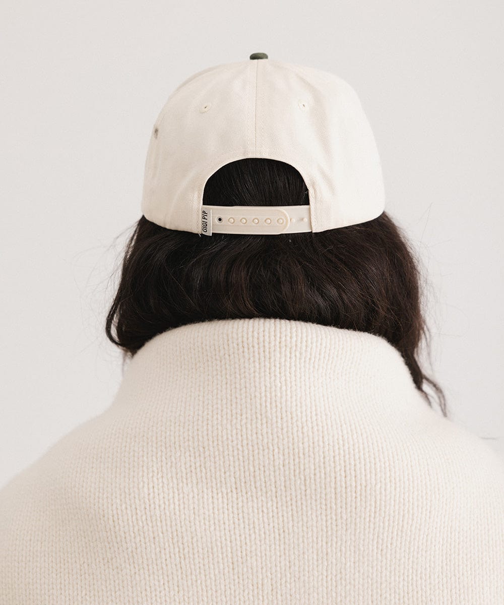 Woman wearing a cream and dark green Trucker hat and a white sweater against a plain background #color_cream - dark green