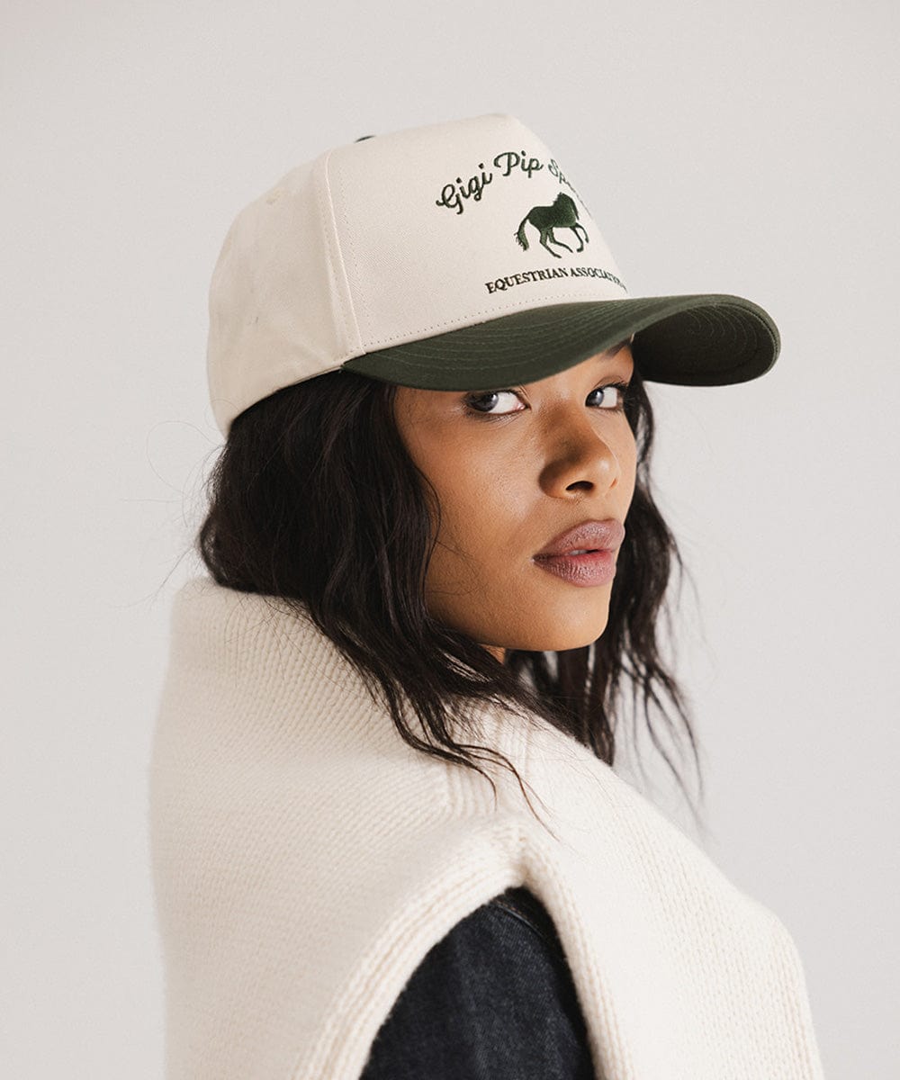 Woman wearing a cream and dark green Trucker hat with 'Gigi Pip Sports' and horse logo and a white sweater, posing sideways against a plain background #color_cream - dark green