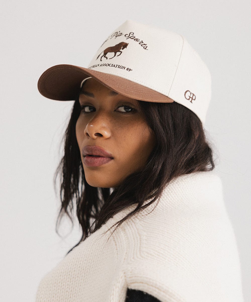 Woman wearing a cream and chocolate brown Trucker hat with 'Gigi Pip Sports' and horse logo and a denim jacket, posing sideways against a plain background #color_cream - chocolate brown
