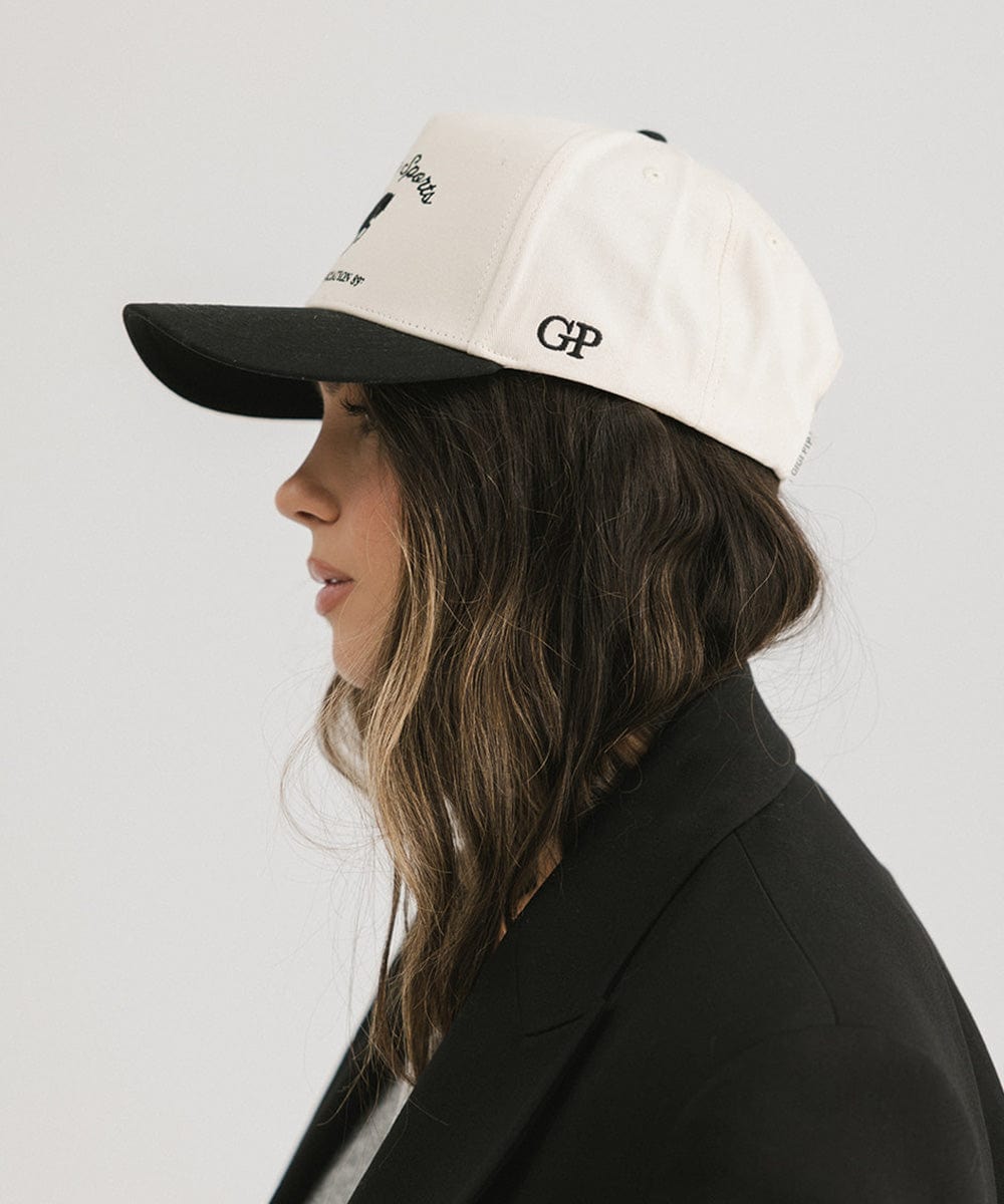 Woman wearing a cream and black Trucker hat with 'Gigi Pip Sports' and horse logo and a black blazer, facing sideways against a plain background #color_cream - black