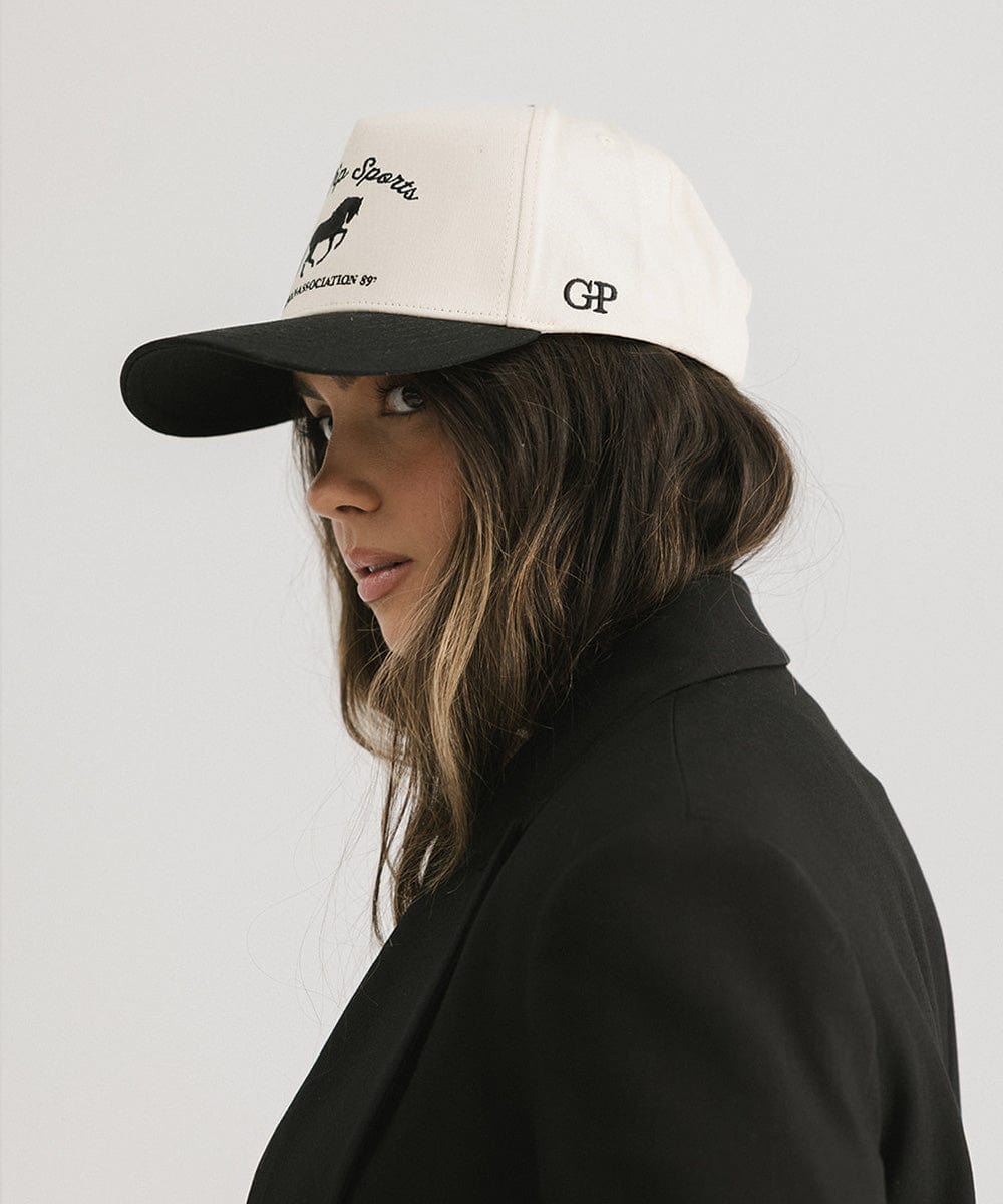 Woman wearing a cream and black Trucker hat with 'Gigi Pip Sports' and horse logo and a black blazer, posing sideways against a plain background #color_cream - black