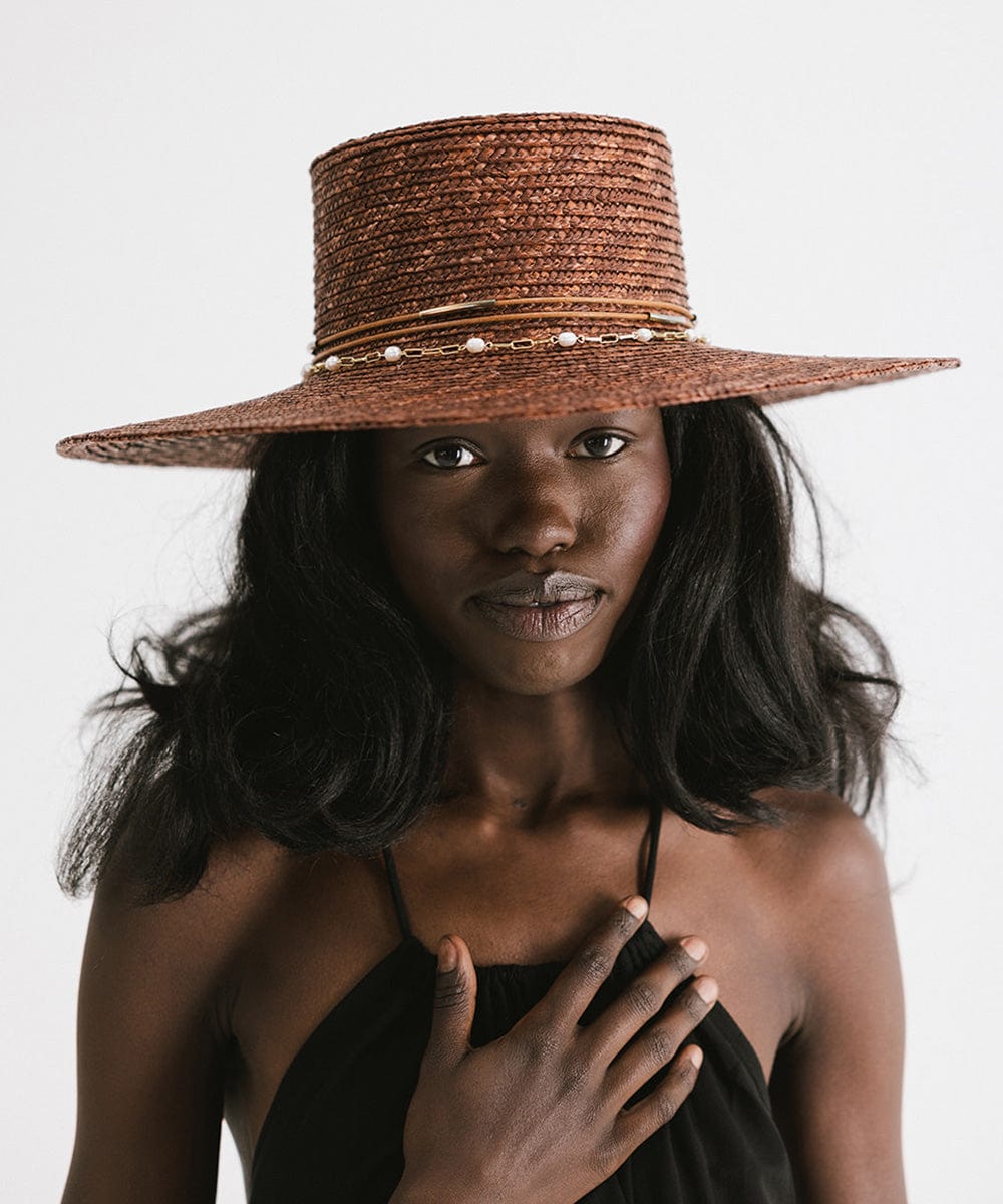 Woman wearing a brown straw hat with a gold pearl paperclip chain band and a black top against a plain background #color_gold