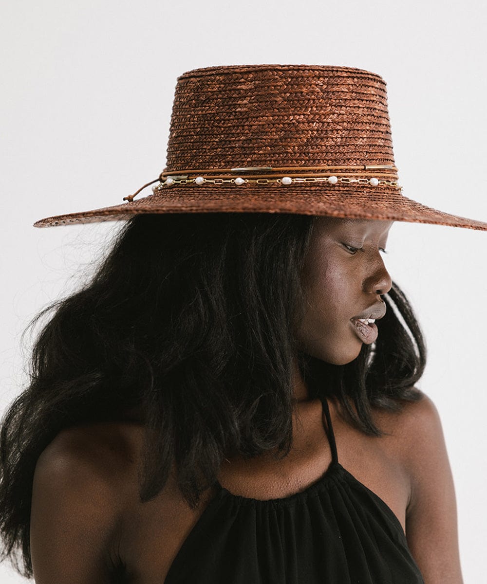 Woman wearing a brown straw hat with a gold pearl paperclip chain band and a black top, looking sideways against a plain background #color_gold