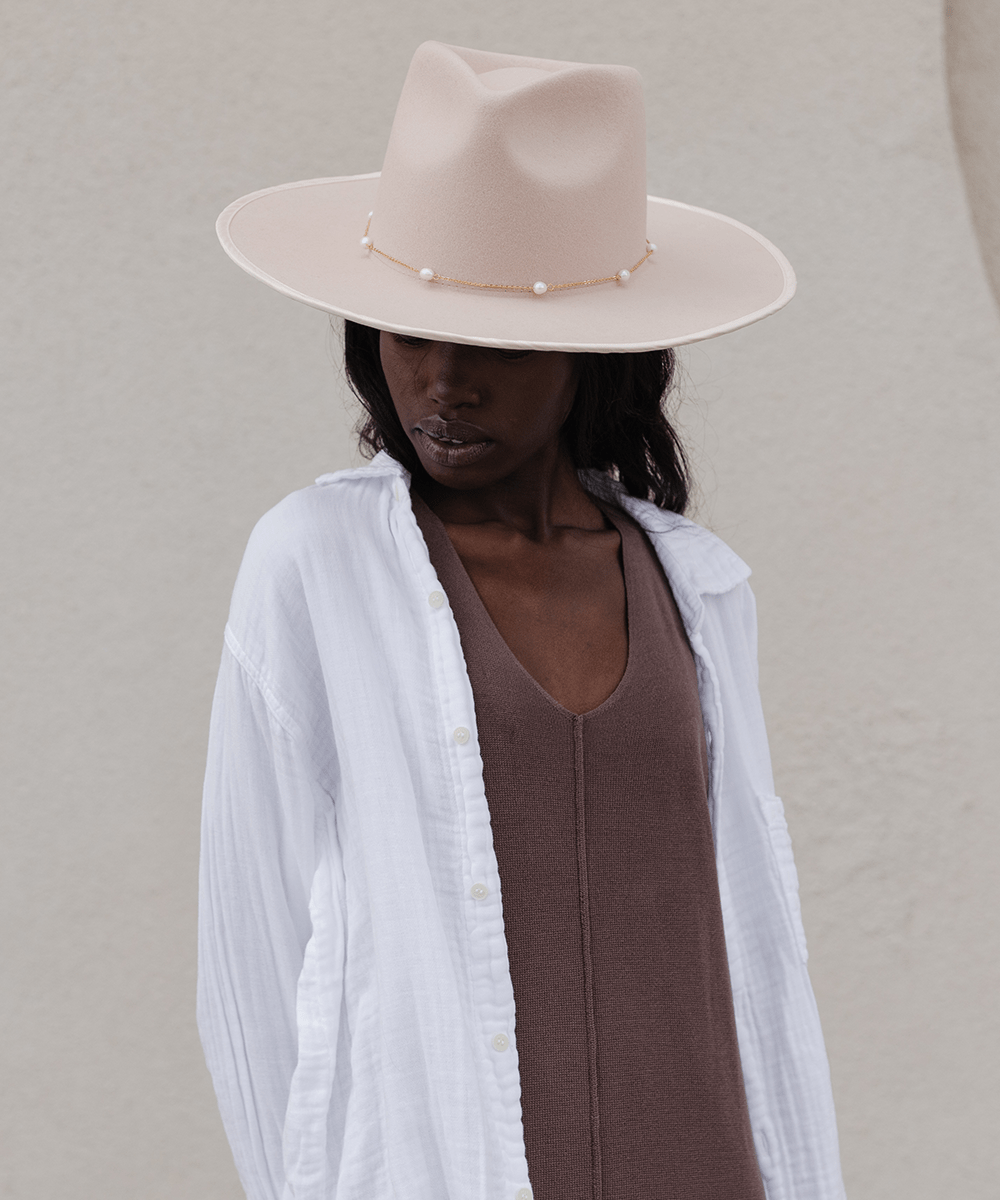 Woman wearing a cream hat with a gold pearl chain band and a brown top with a white shirt against a plain background #color_gold