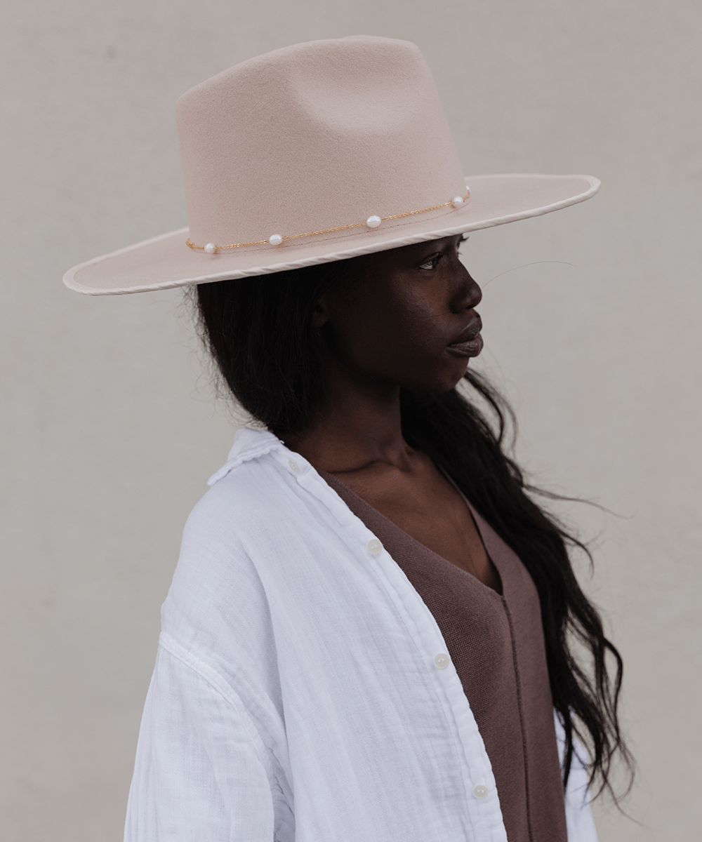 Woman wearing a cream hat with a gold pearl chain band and a brown top with a white shirt, facing sideways against a plain background #color_gold