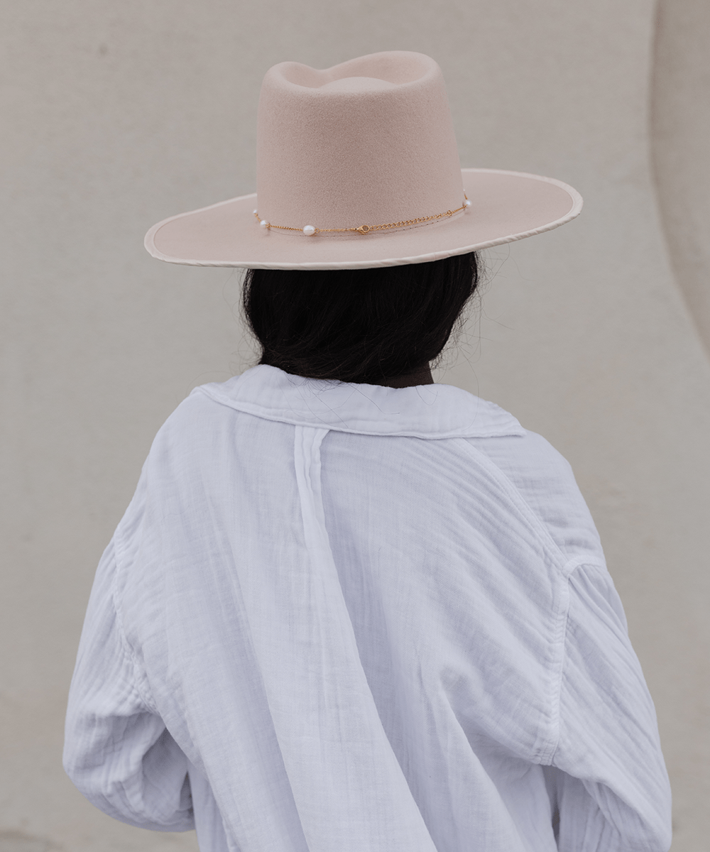 Woman wearing a cream hat with a gold pearl chain band and a white shirt, facing behind against a plain background #color_gold