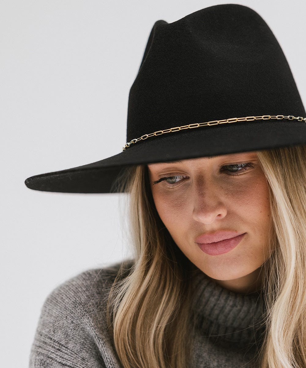 Close up image of a woman wearing a black wide-brimmed hat with a gold paperclip chain hat band against a plain background #color_gold