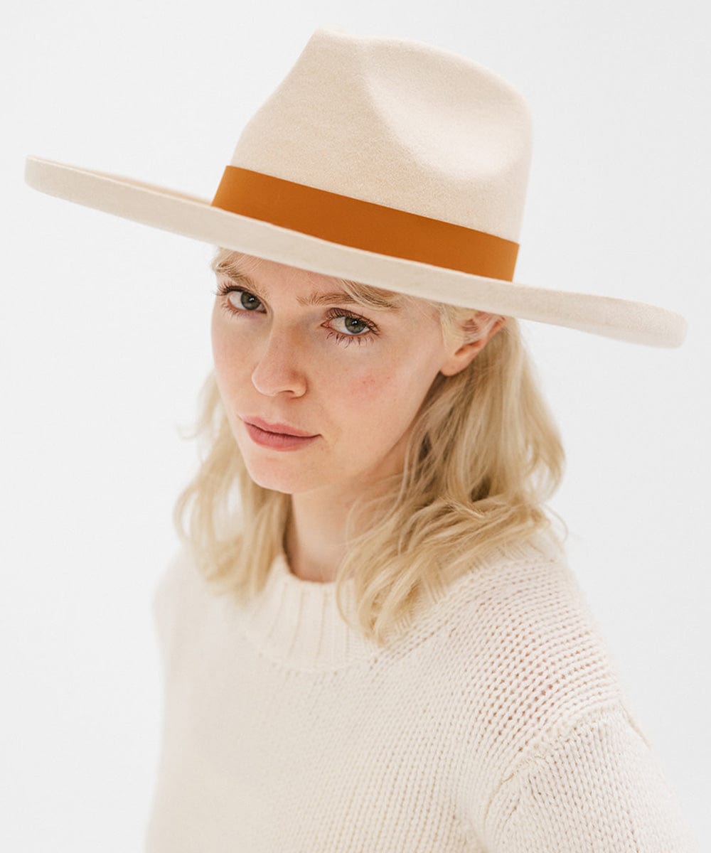 A woman wearing a cream hat with a wide cognac leather band around the base, and a cream sweater against a plain background #color_cognac