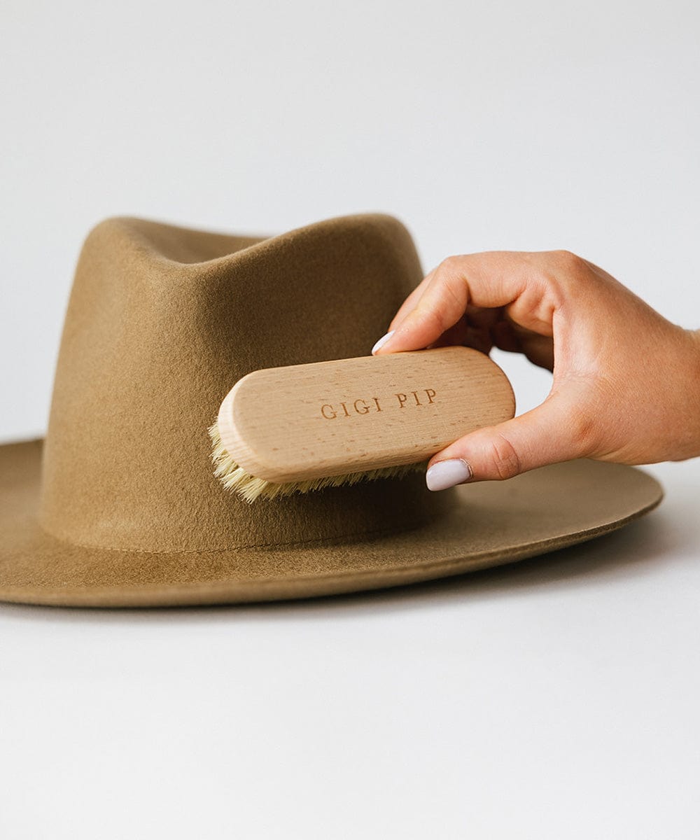 Person holding a cream colored hat brush against a brown hat on a plain background