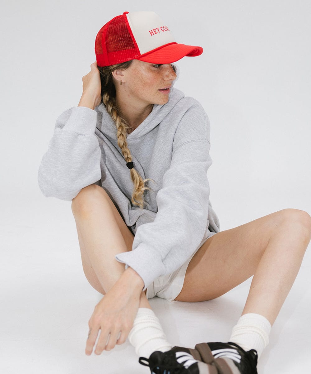 Woman wearing a cream and vintage red cap with 'Hey Cowboy' text and a grey sweater, sitting against a plain background #color_cream-vintage red