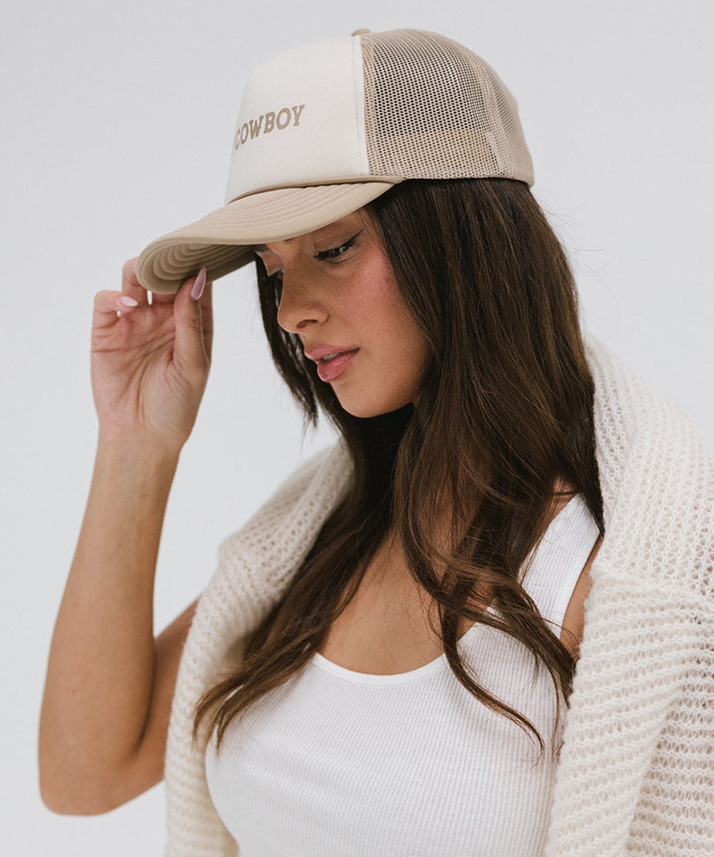 Woman wearing a tan and cream cap with 'Hey Cowboy' text and a white top, posing sideways against a plain background #color_tan-cream
