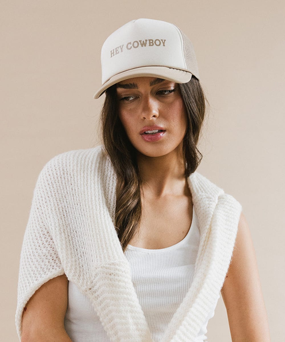 Woman wearing a tan and cream cap with 'Hey Cowboy' text and a white top with a white sweater against a plain background #color_tan-cream