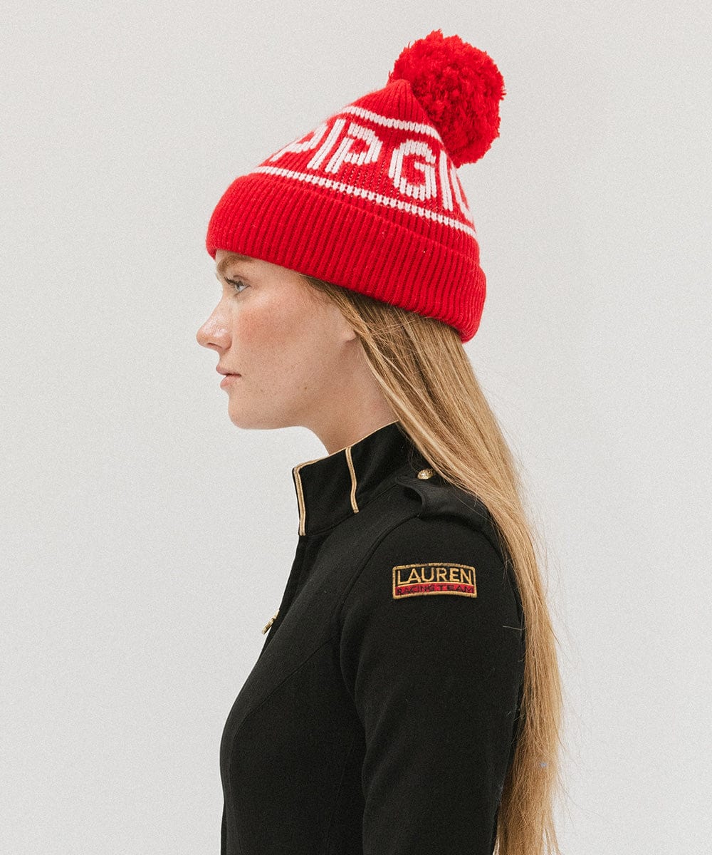 Woman wearing a ruby red colored knit beanie with pom-pom and 'GIGI PIP' branding and a black jacket, facing sideways against a plain background #color_ruby red