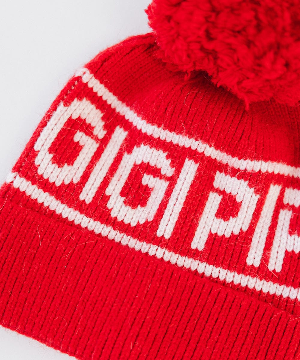 Close up image of a ruby red colored knit beanie with pom-pom and 'GIGI PIP' branding on a white background #color_ruby red
