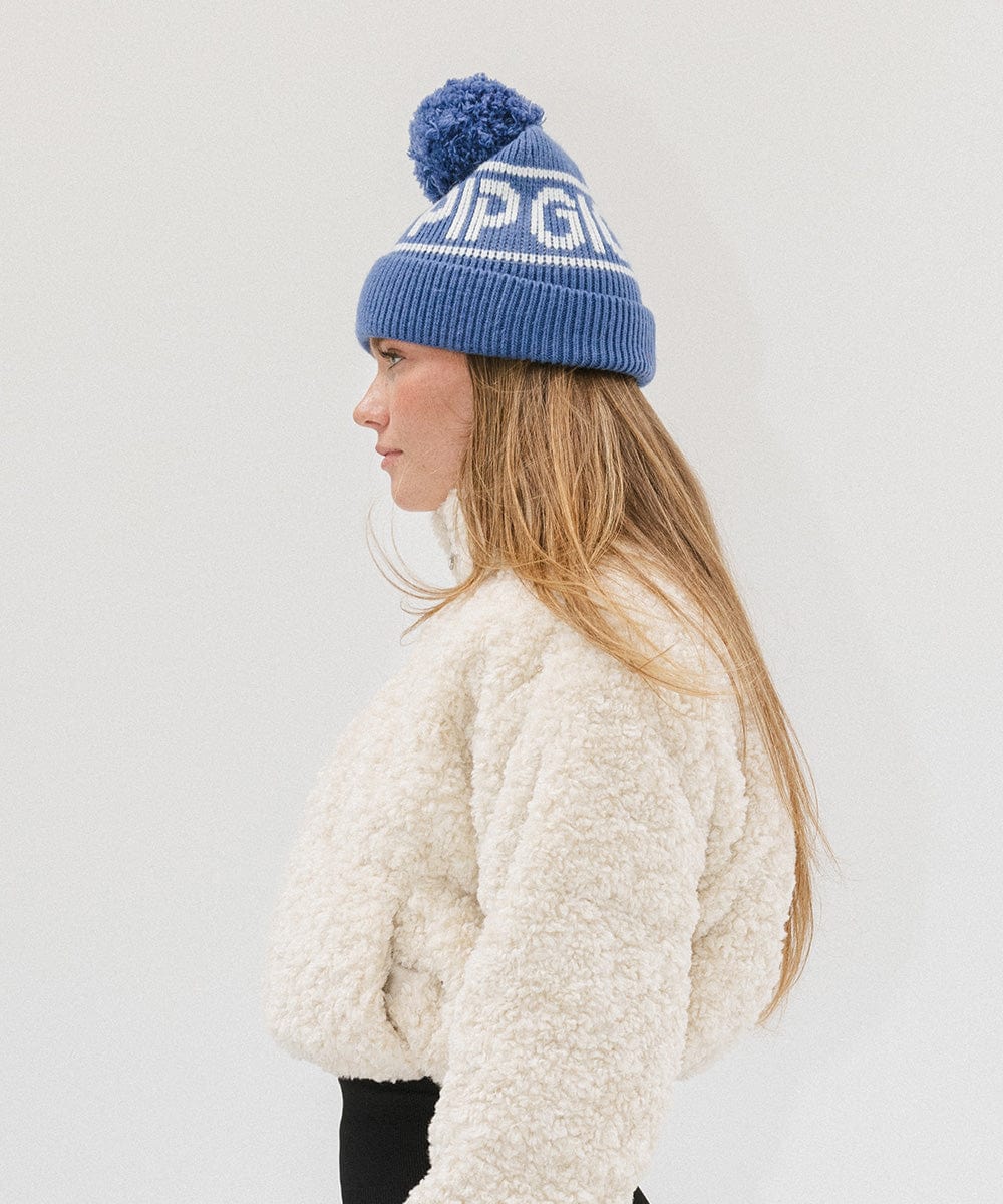 Woman wearing an arctic blue colored knit beanie with pom-pom and 'GIGI PIP' branding and a white sweater, facing sideways against a plain background #color_arctic blue