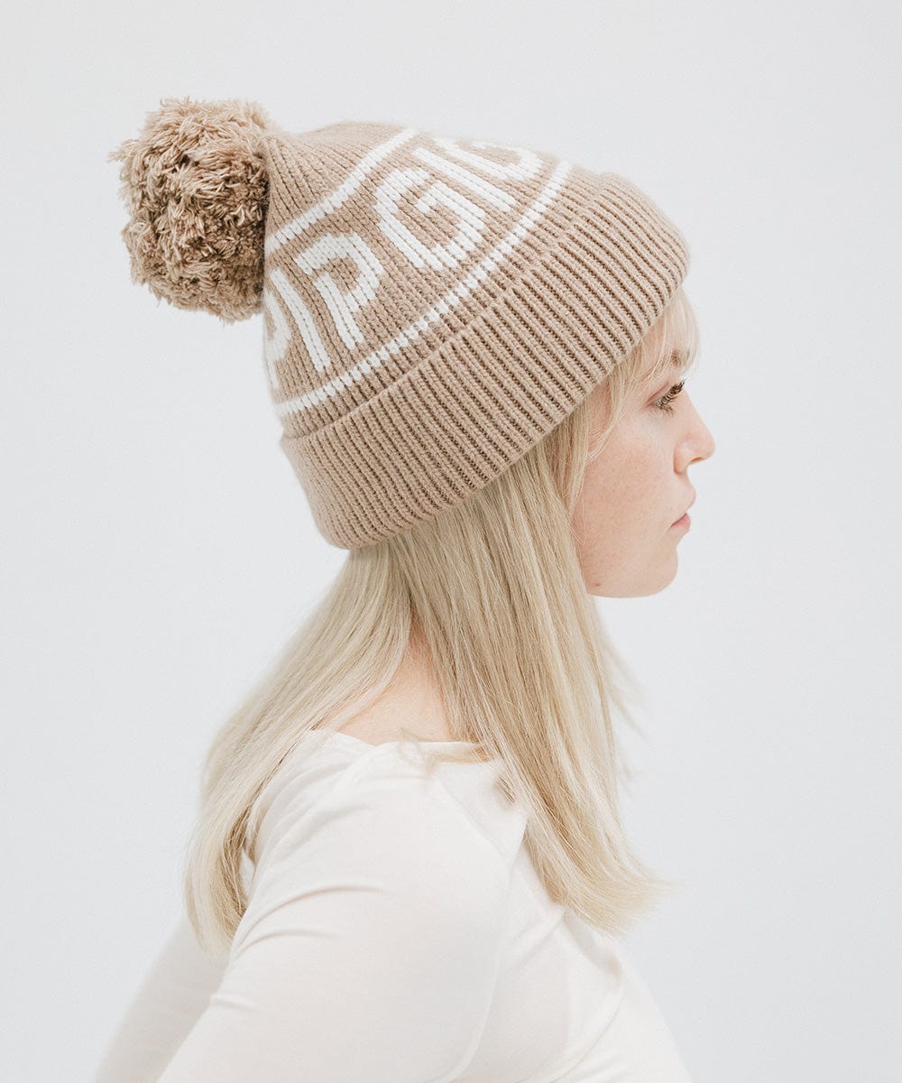 Woman wearing a taupe colored knit beanie with pom-pom and 'GIGI PIP' branding and a white top, facing sideways against a plain background #color_taupe
