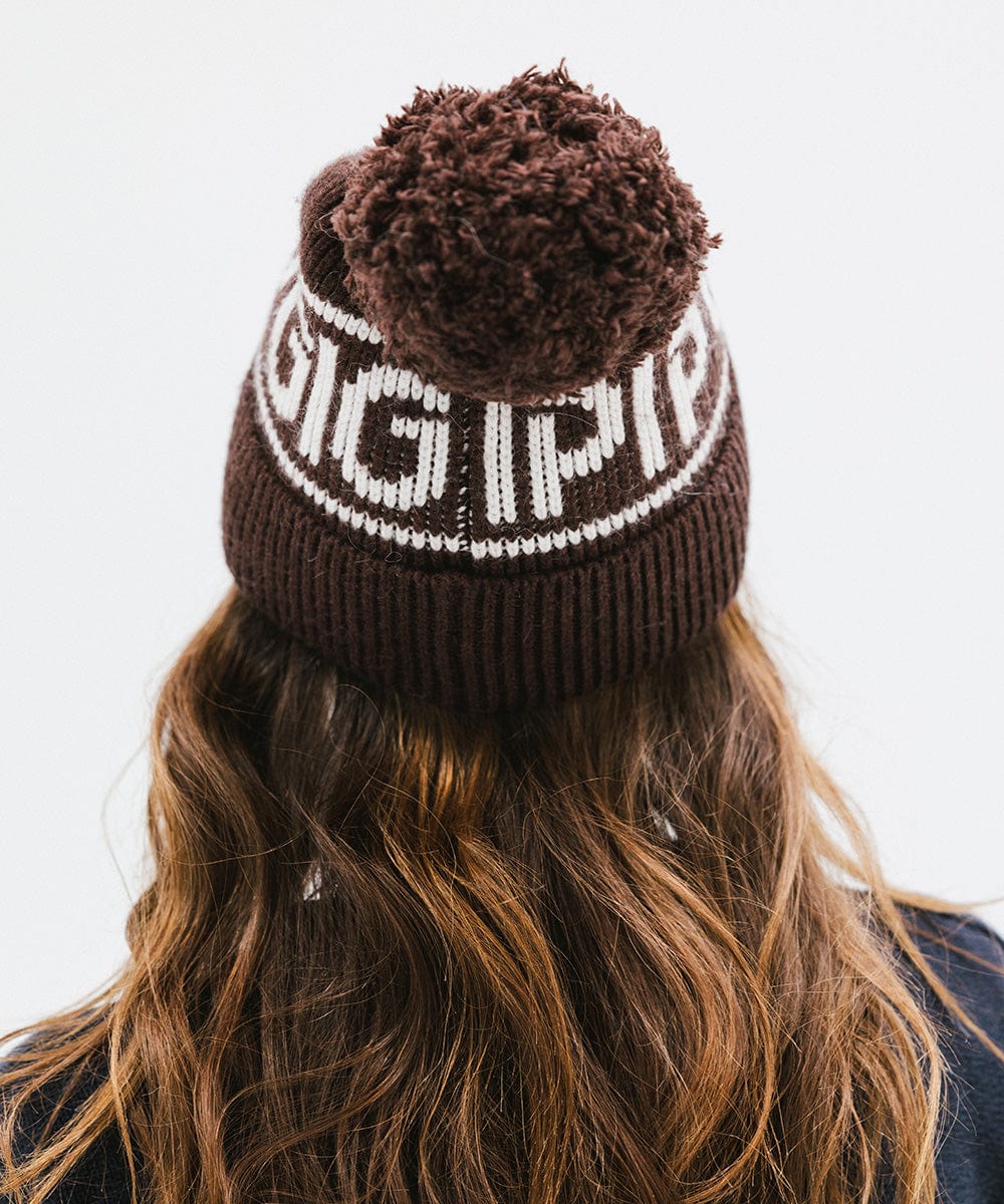 Woman wearing a chocolate brown colored knit beanie with pom-pom and 'GIGI PIP' branding and a dark blue sweater, facing behind against a plain background #color_chocolate brown