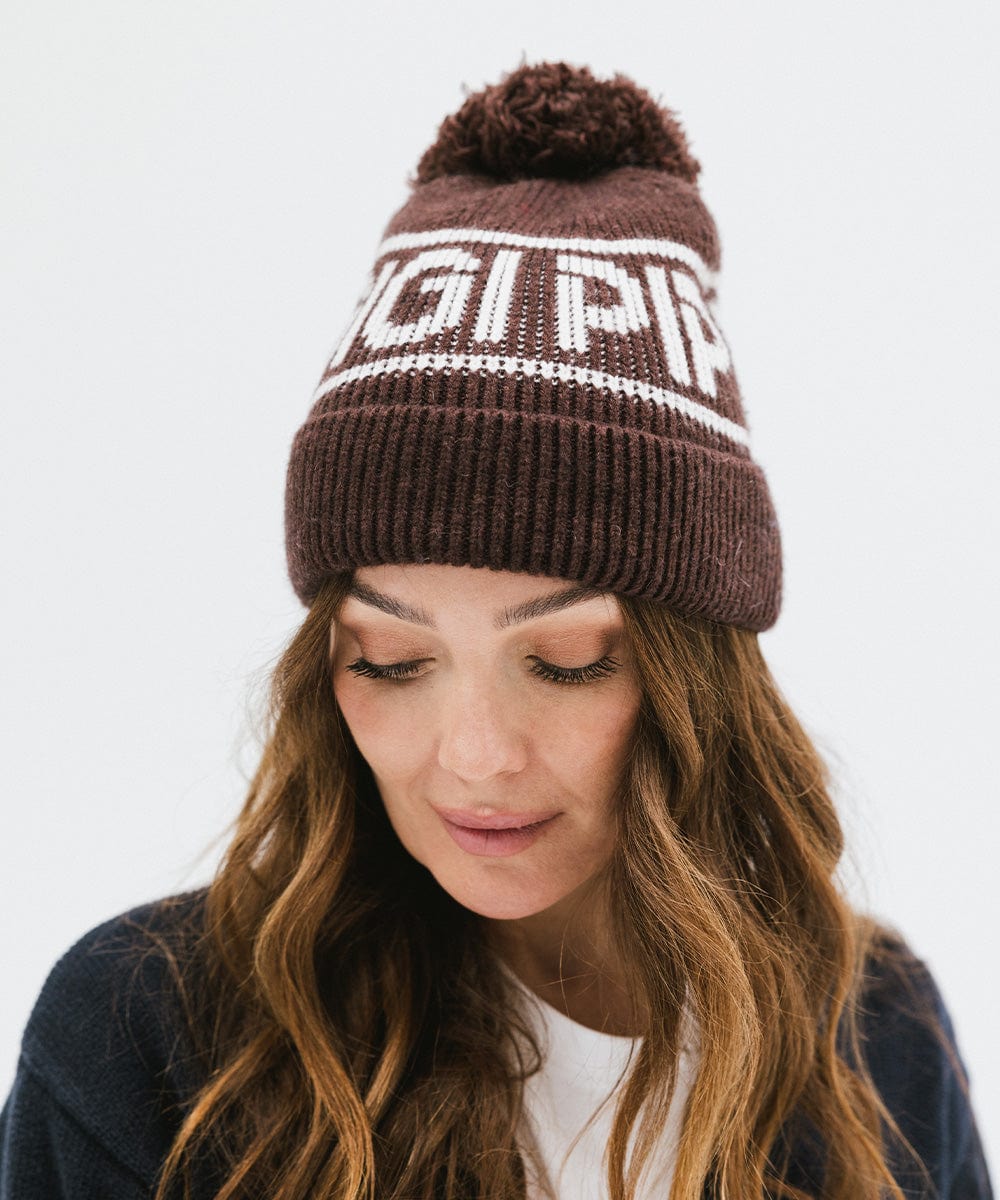 Woman wearing a chocolate brown colored knit beanie with pom-pom and 'GIGI PIP' branding and a dark blue sweater against a plain background #color_chocolate brown