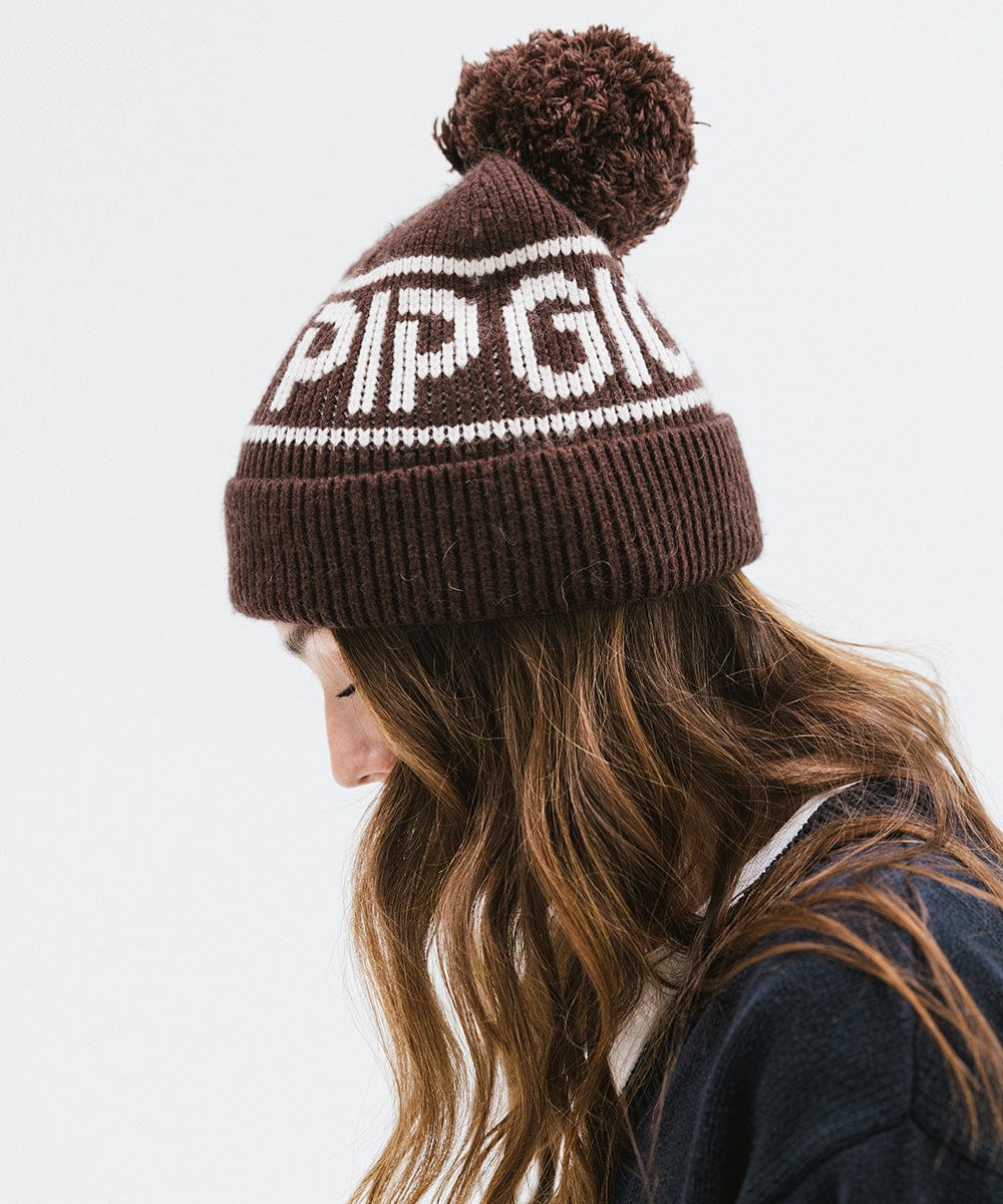 Woman wearing a chocolate brown colored knit beanie with pom-pom and 'GIGI PIP' branding and a dark blue sweater, facing sideways against a plain background #color_chocolate brown