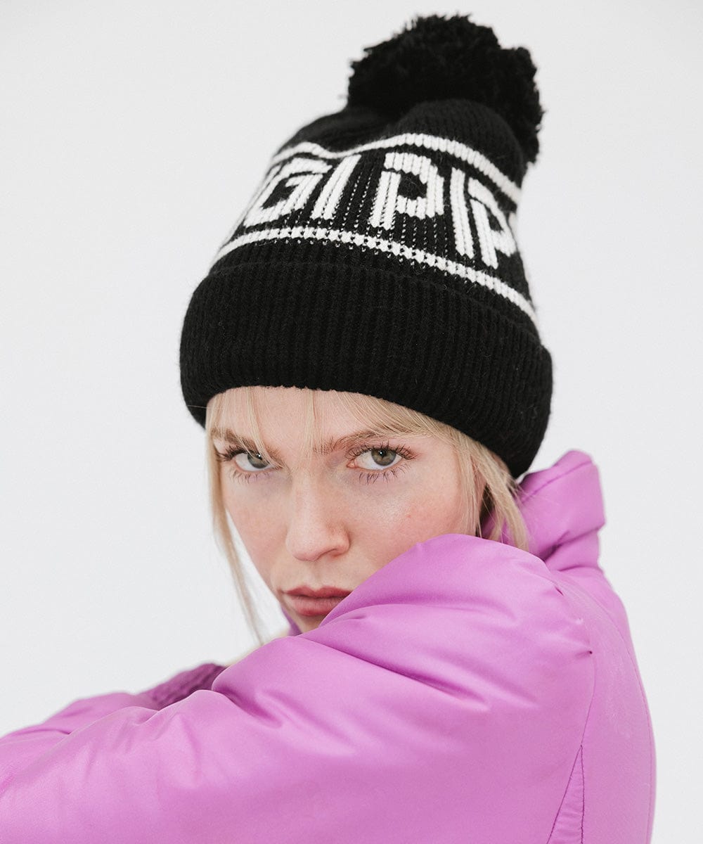Woman wearing a black knit beanie with pom-pom and 'GIGI PIP' branding and a purple jacket against a plain background #color_black