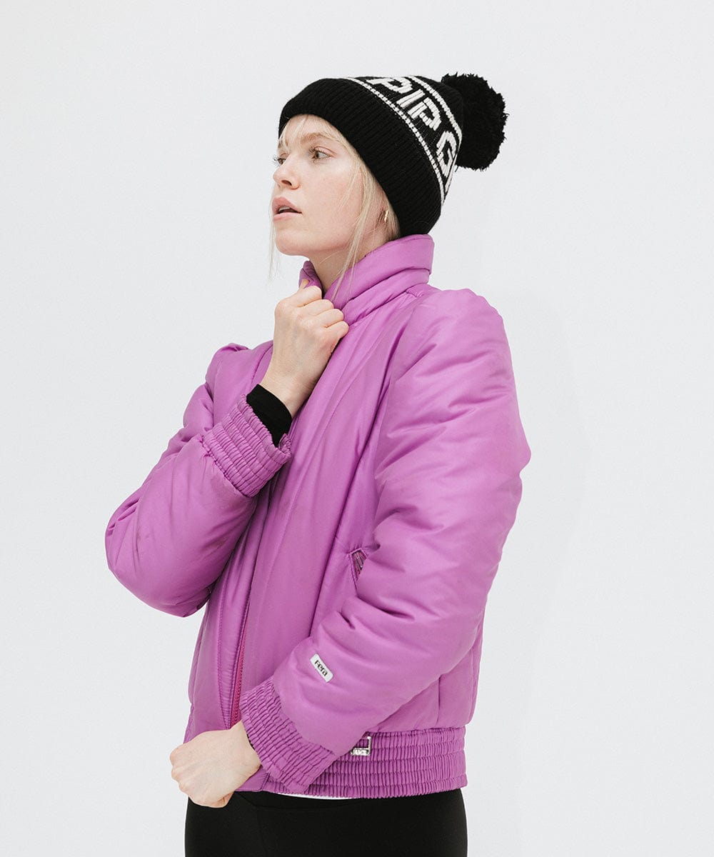 Woman wearing a black knit beanie with pom-pom and 'GIGI PIP' branding and a purple jacket, posing sideways against a plain background #color_black