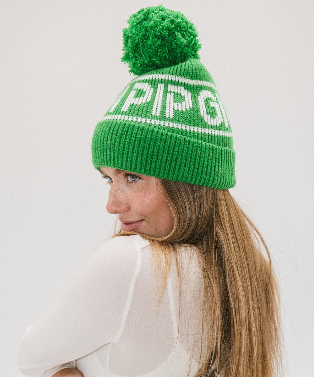 Woman wearing an evergreen colored knit beanie with pom-pom and 'GIGI PIP' branding and a white top, facing sideways against a plain background #color_evergreen