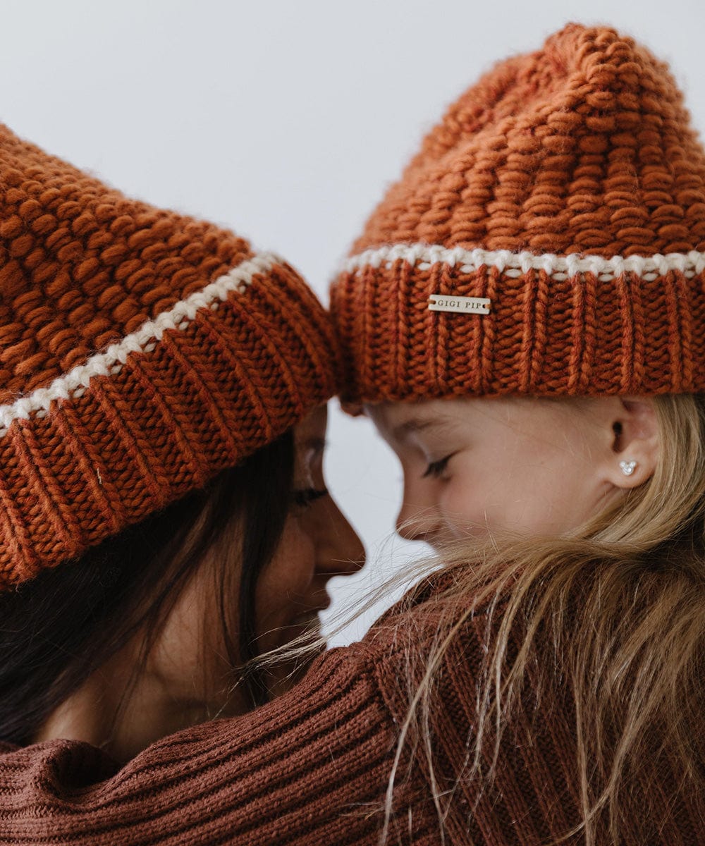 A woman and a child, both wearing a burnt orange vail beanie with a cream stripe facing each other against a plain background #color_burnt orange-cream