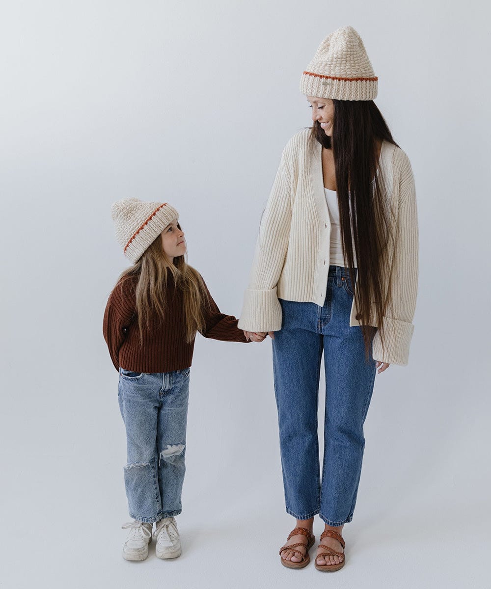 Child wearing a cream vail beanie with a burnt orange stripe, a brown sweater, blue jeans and white shoes with a woman wearing a cream vail beanie with a burnt orange stripe, a cream jacket, blue jeans and brown sandals, both looking at each other while holding hands against a plain background #color_cream-burnt orange