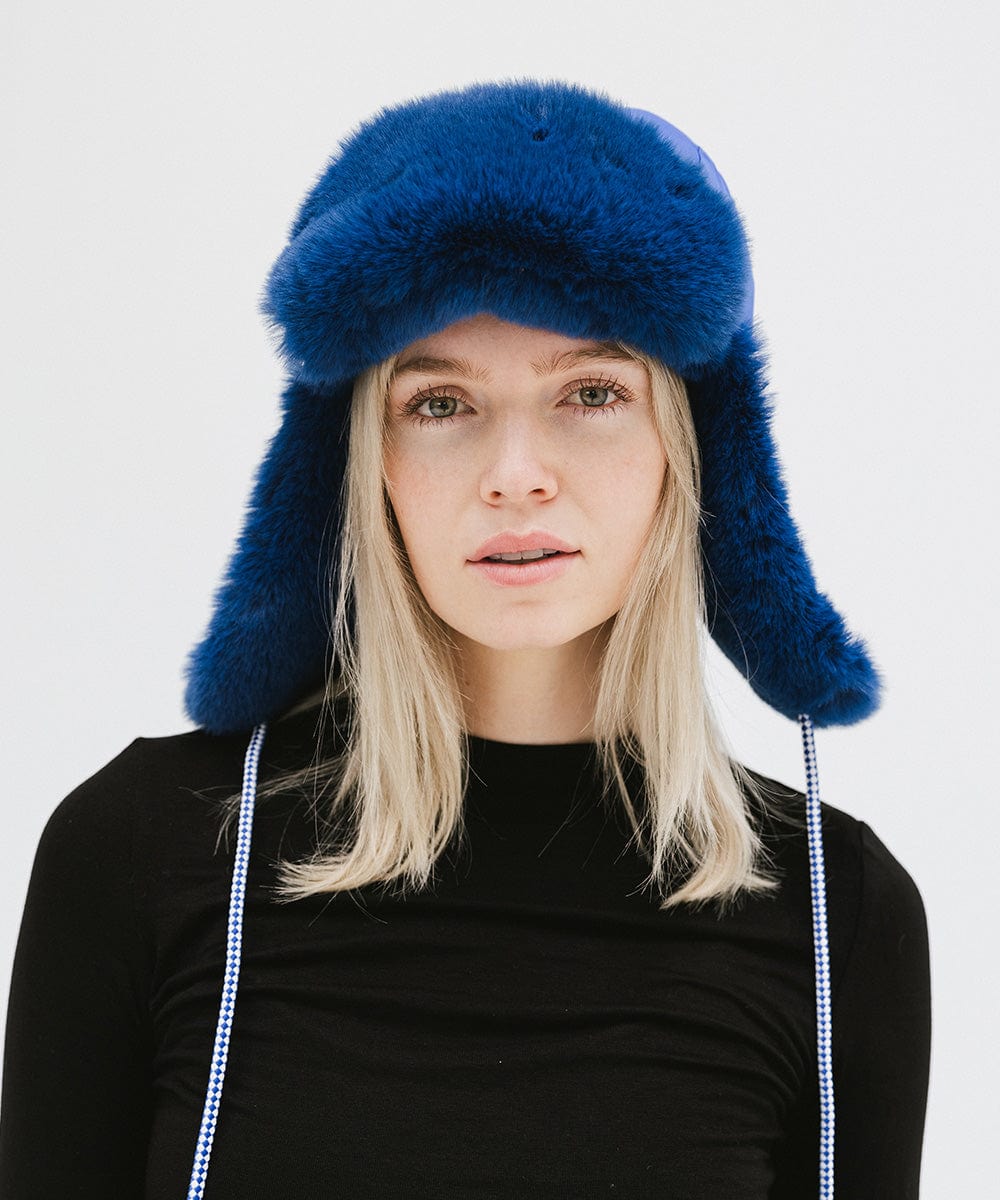 Woman wearing an alpine blue colored fur-trimmed hat and a black top against a plain background #color_alpine blue