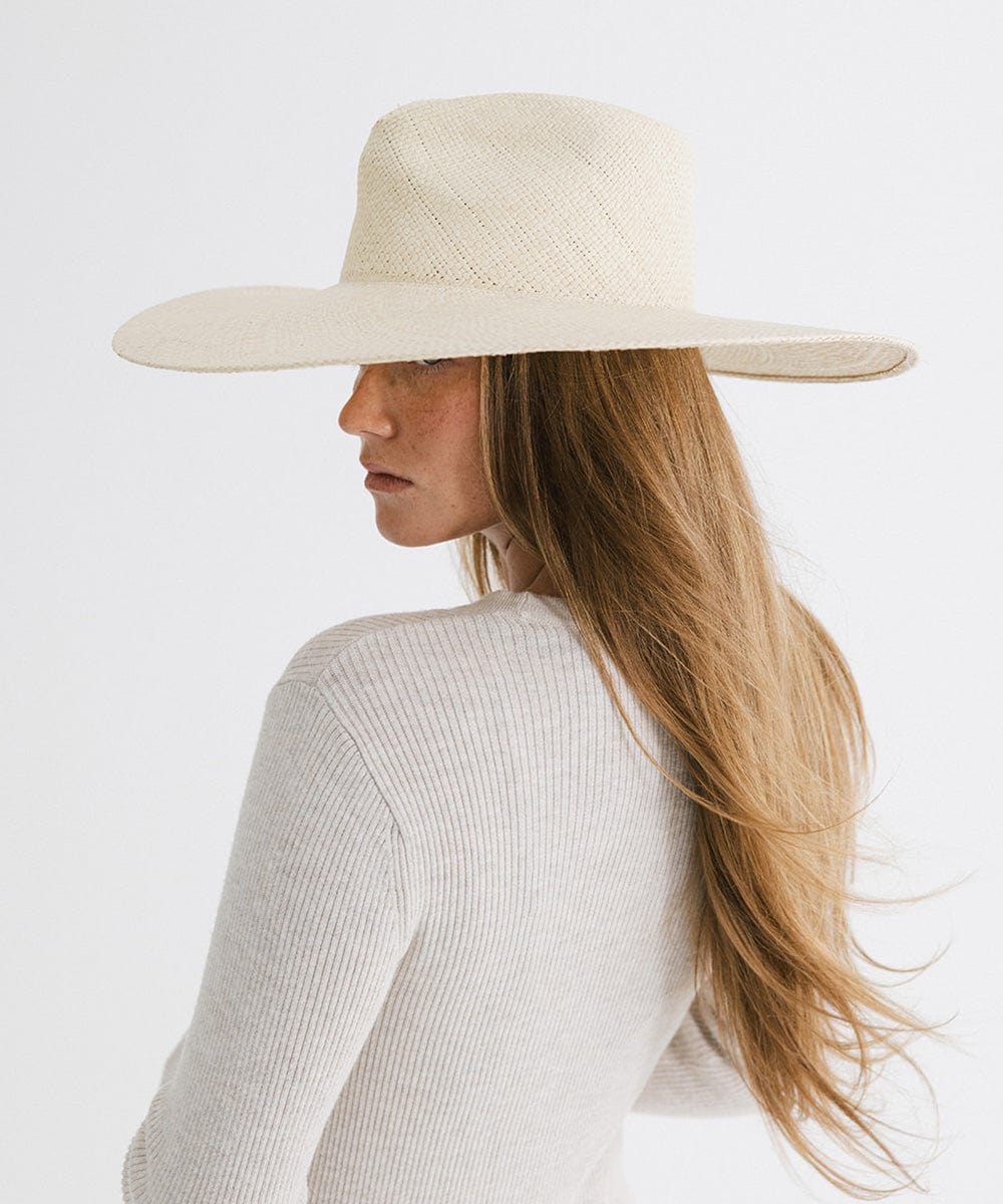 Woman with long hair wearing a natural colored wide brim straw hat and a white top, posing sideways against a plain background #color_natural