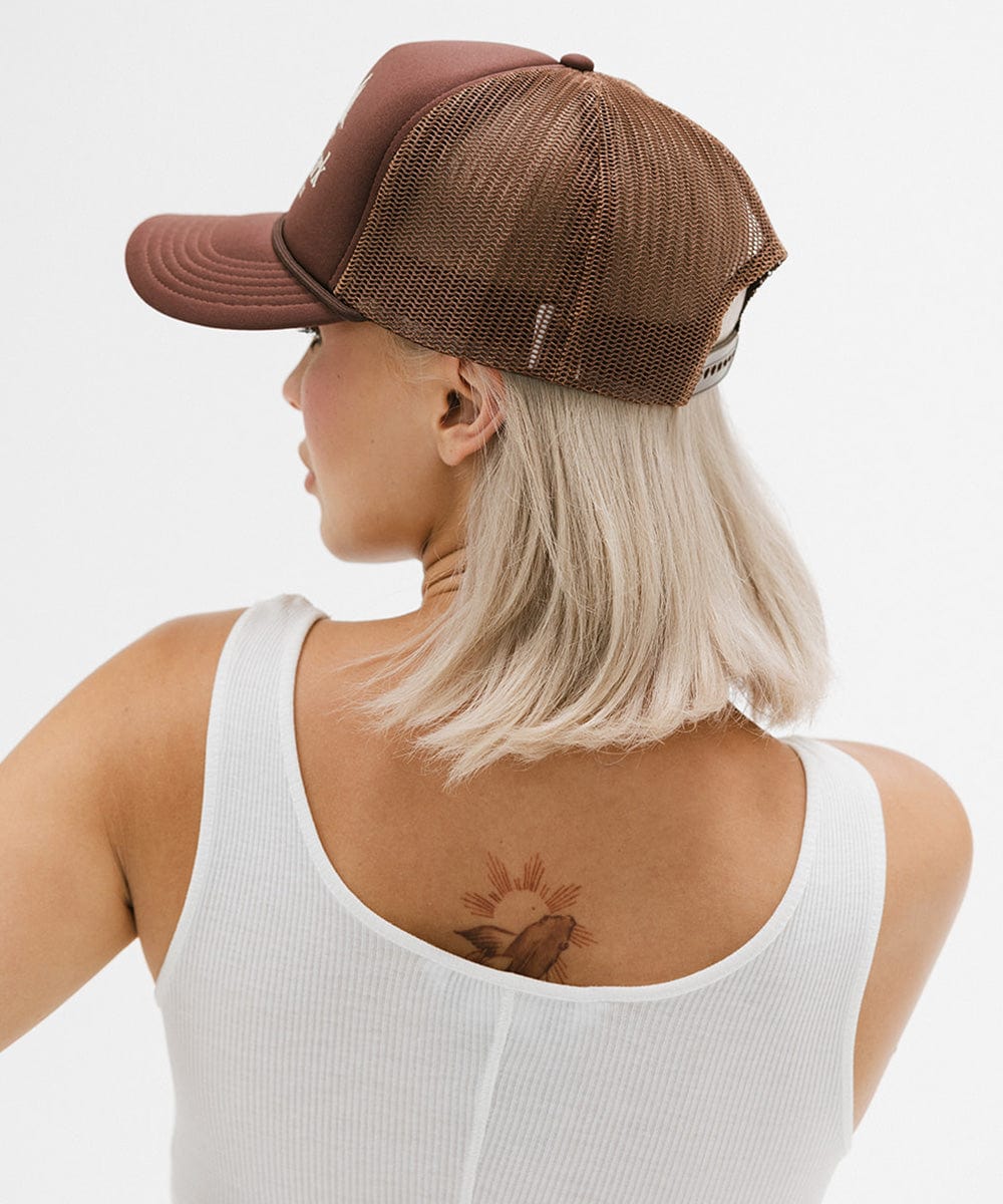 Woman wearing a brown trucker hat and a white top, facing sideways against a plain background #color_chocolate brown