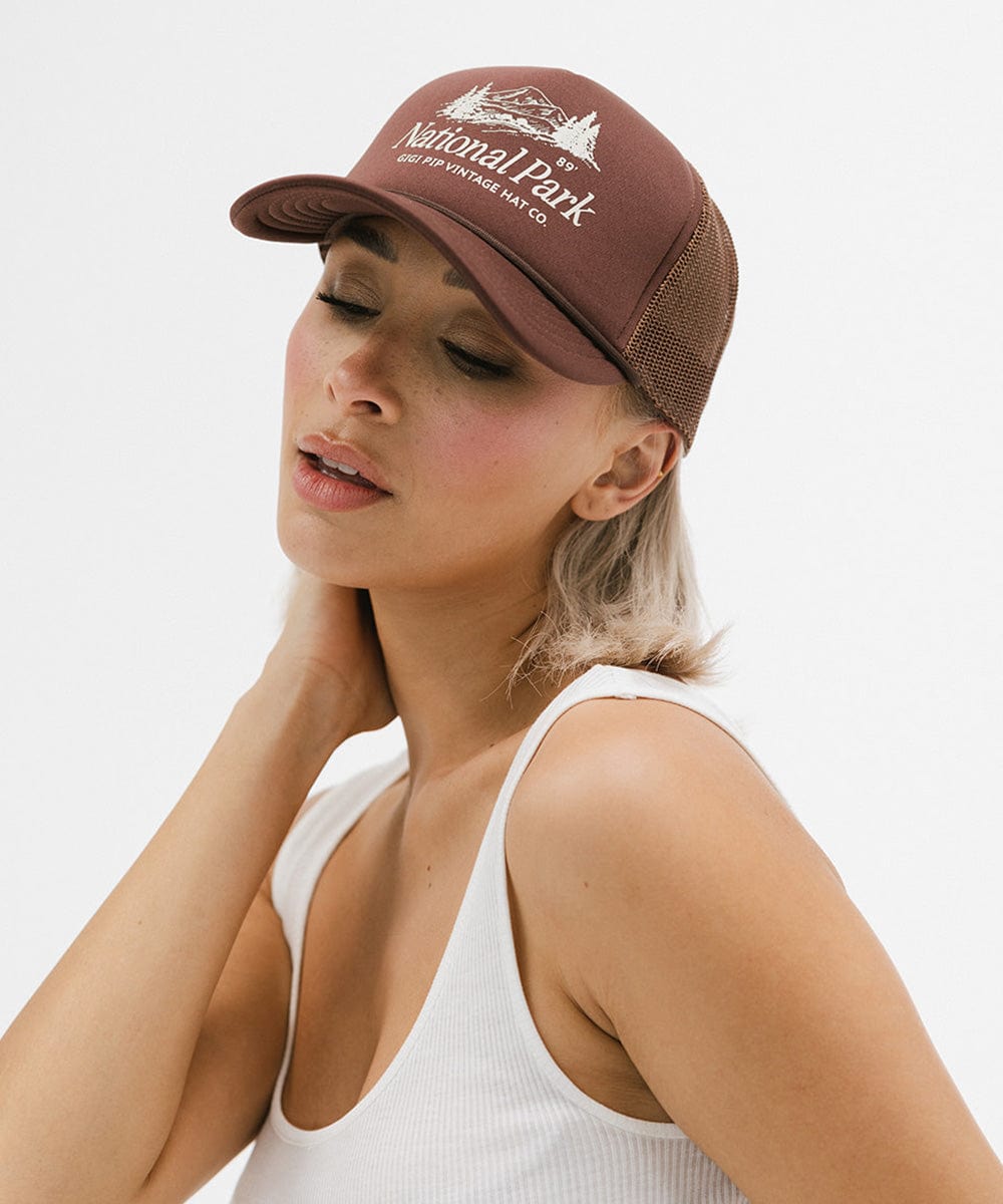 Woman wearing a brown trucker hat with 'National Park' logo and a white top, posing against a plain background #color_chocolate brown