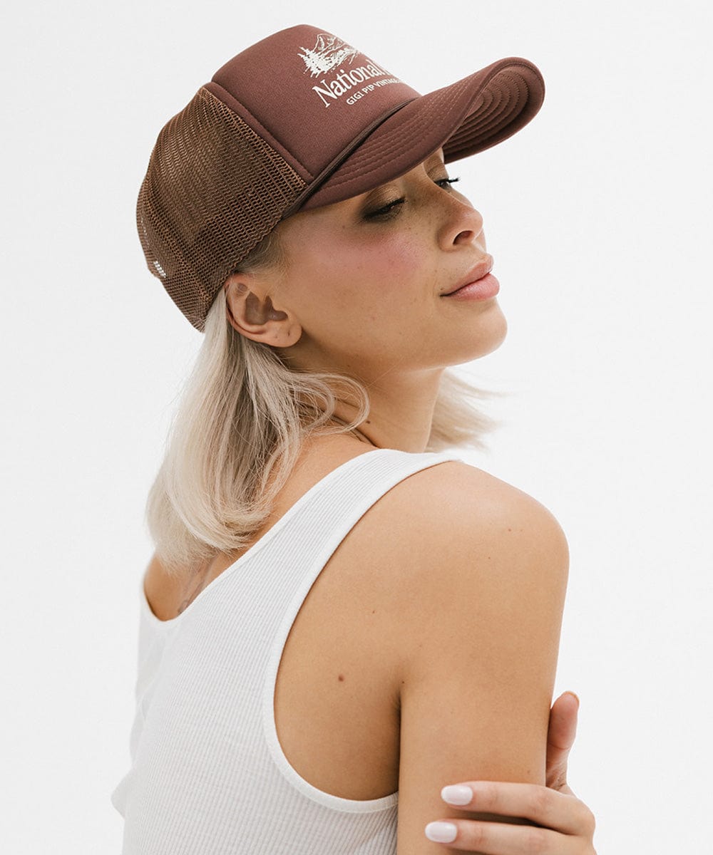 Woman wearing a brown trucker hat with 'National Park' logo and a white top, posing sideways against a plain background #color_chocolate brown