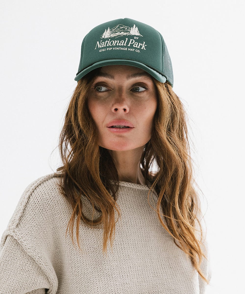 Woman wearing a forest green colored trucker hat with 'National Park' logo and a grey sweatshirt against a plain background #color_forest green