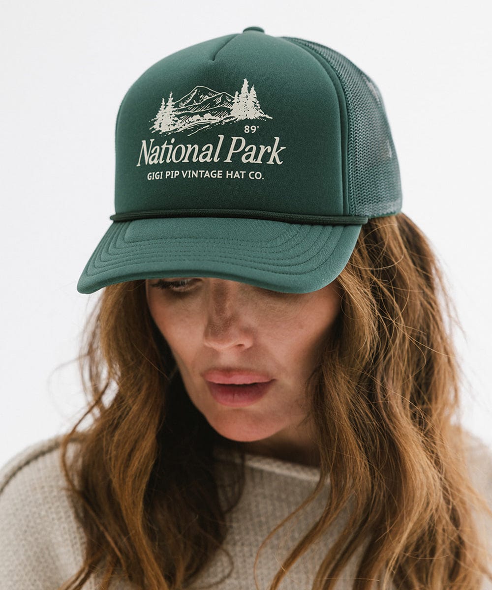 Woman wearing a forest green colored trucker hat with 'National Park' logo against a plain background #color_forest green