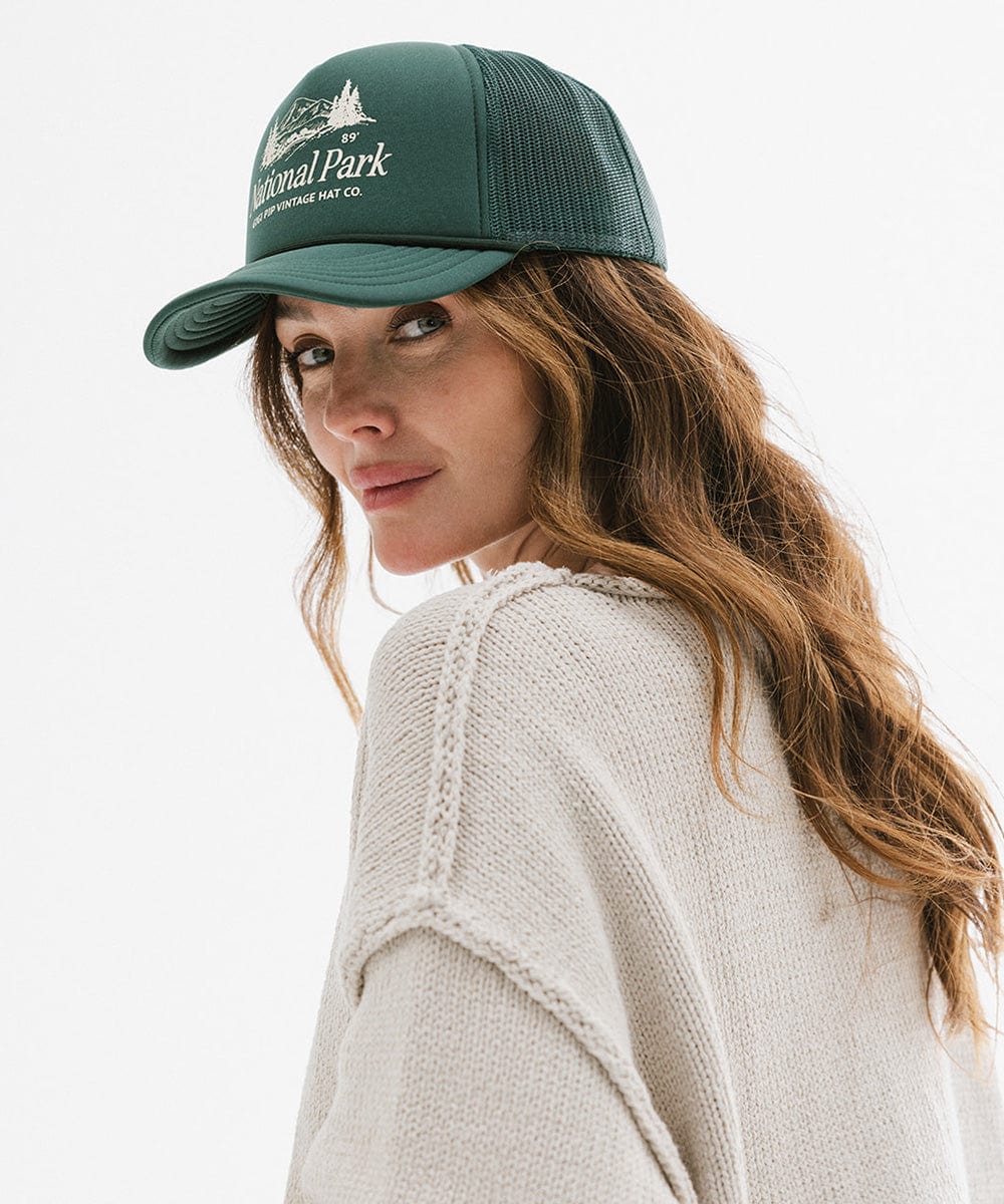 Woman wearing a forest green colored trucker hat with 'National Park' logo and a grey sweatshirt, posing sideways against a plain background #color_forest green