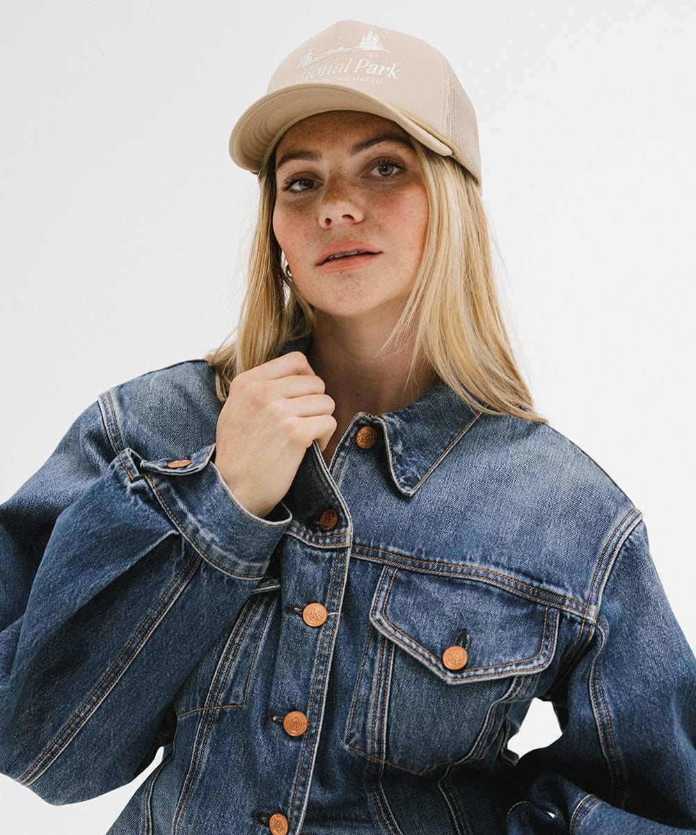 Woman wearing a tan trucker hat with 'National Park' logo and a denim jacket against a plain background #color_tan