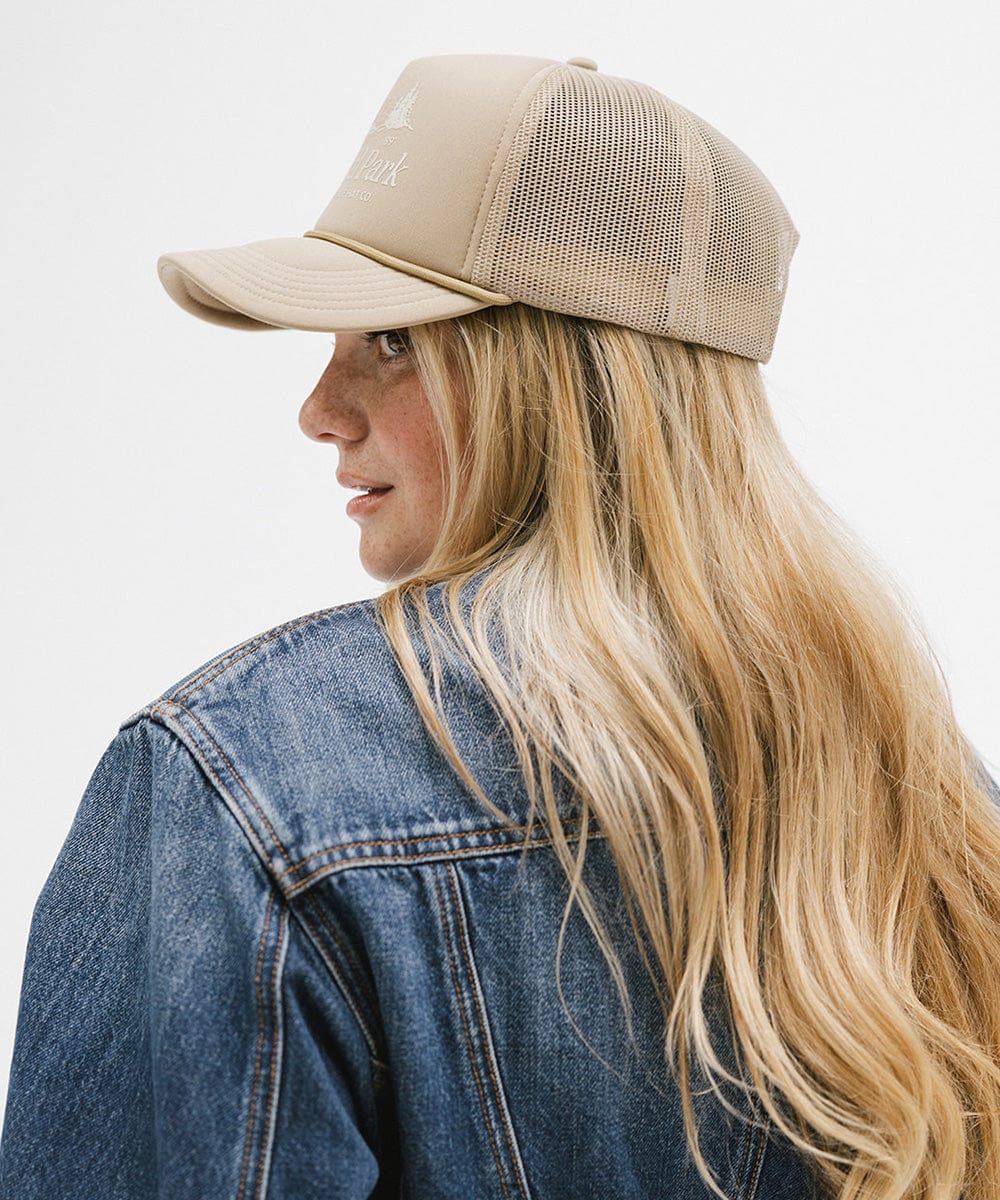 Woman wearing a tan trucker hat with 'National Park' logo and a denim jacket, facing sideways against a plain background #color_tan