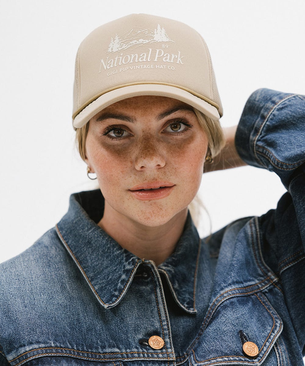 Woman wearing a tan trucker hat with 'National Park' logo and a denim jacket against a plain background #color_tan
