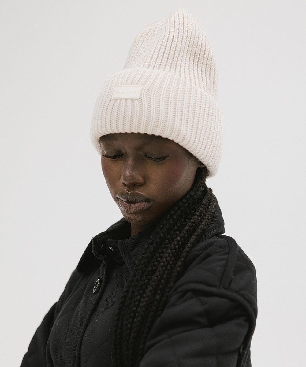 A woman wearing an off white colored ribbed beanie and a black jacket against a plain background #color_off white