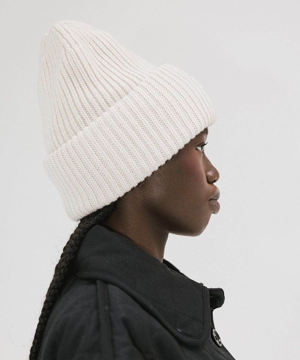 A person wearing an off white colored ribbed beanie, facing sideways against a plain background #color_off white