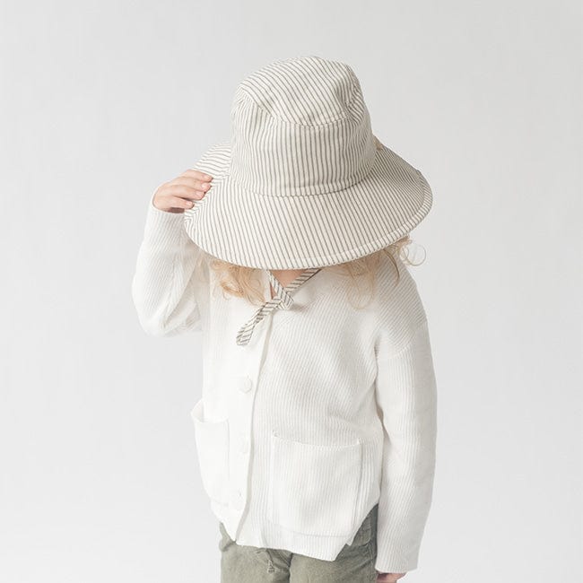 Young girl wearing a grey striped bucket hat and a white shirt with grey pants, looking down against a plain background #color_grey