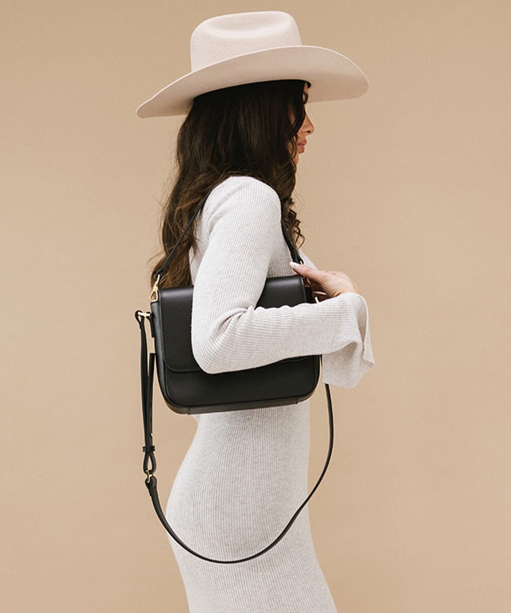 Woman wearing a hat and a white outfit, carrying a black Rhys Crossbody Genuine Leather Bag, facing sideways against a plain background #color_black