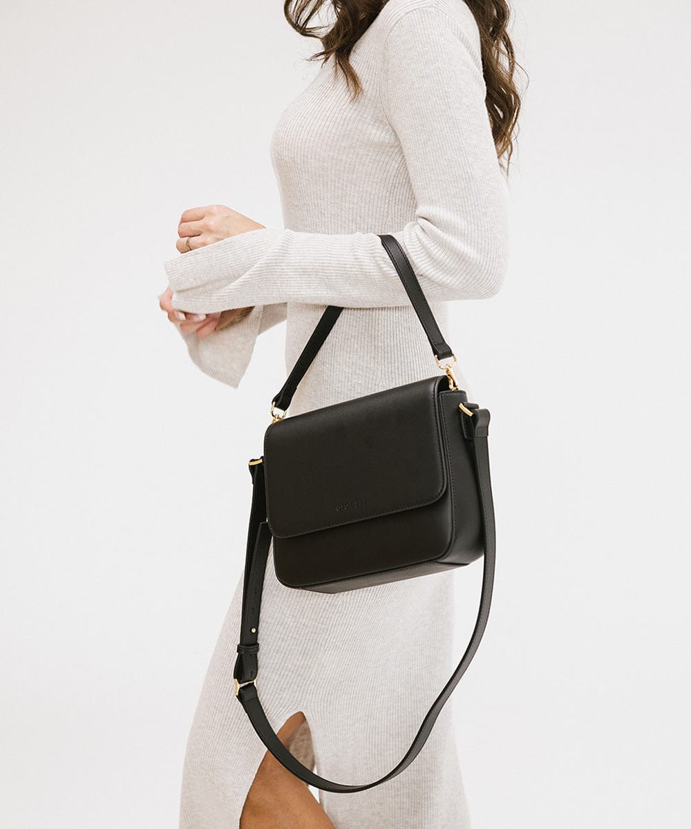 Person wearing a white outfit carrying a black Rhys Crossbody Genuine Leather Bag, posing sideways against a plain background #color_black