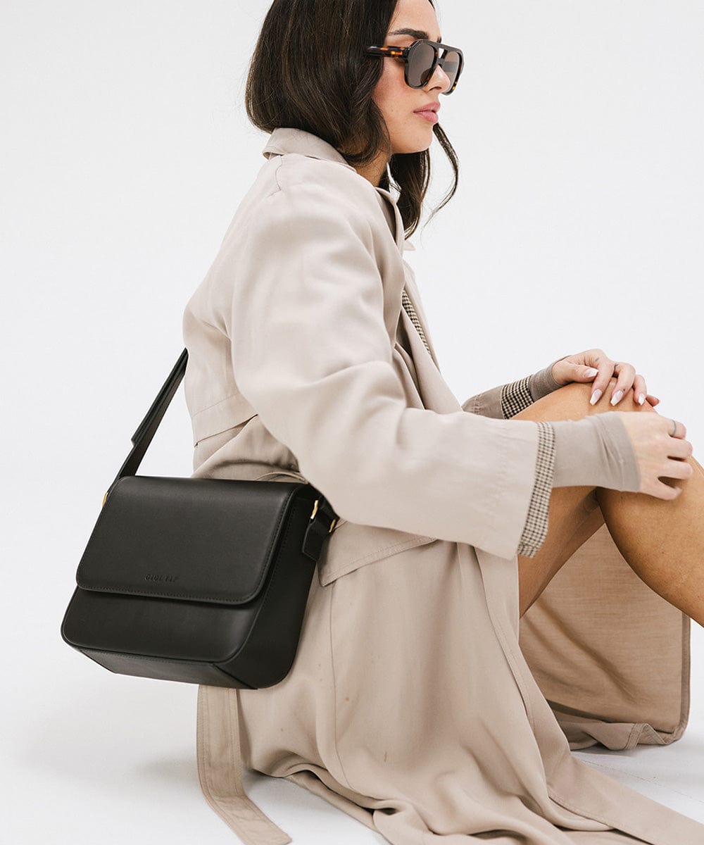 Woman wearing sunglasses and a beige coat, carrying a black Rhys Crossbody Genuine Leather Bag, sitting against a plain background #color_black